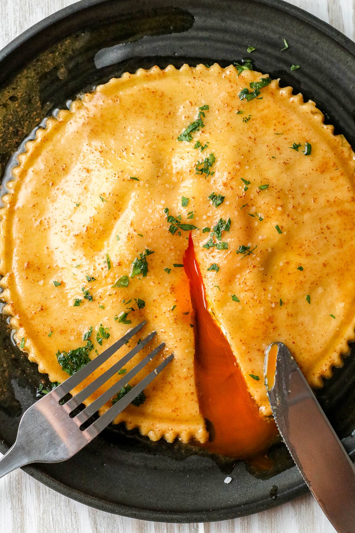 knife and fork cutting into raviolo on a black plate.